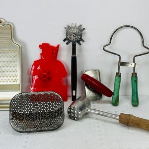 May include: A collection of vintage kitchen tools including a red plastic cookie cutter shaped like a person, a metal grater, a metal can opener, a metal meat tenderizer with a wooden handle, a metal grill press with a black handle, and a metal wire frame with two green handles.