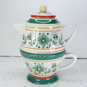 May include: A ceramic tea for one set featuring a teapot stacked on a teacup. The cream-colored ceramic is decorated with green floral patterns, teal speckled bands, and thin red stripes. The teapot has a small yellow knob on the lid.