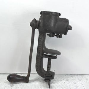 May include: An antique, cast iron food chopper with a hand crank. The vintage kitchen tool is dark gray and features a clamp for securing it to a table. The handle and crank are made of wood.