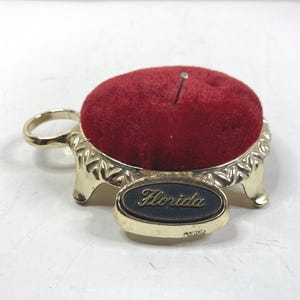 May include: A vintage pin cushion with a red velvet top and a gold-colored metal base. The base has a decorative design and a small oval plaque that reads "Florida". A single pin is inserted into the cushion.
