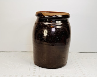 May include: A cylindrical stoneware crock featuring a dark brown, high-gloss glaze that covers the exterior body. The rim is unglazed, revealing a natural tan clay color. The vessel has a wide opening and a sturdy, flat base for stability.
