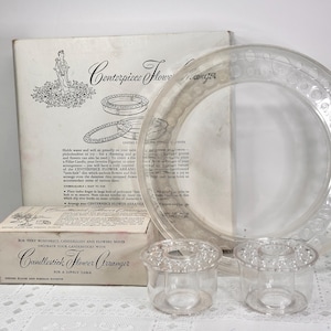 May include: A clear, vintage centerpiece flower arranger set. Includes a large circular plate with decorative holes, two small candle holders, and a box with the text "Candlestick Flower Arranger".