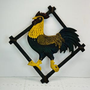 May include: A cast iron rooster wall decor piece. The rooster is painted with yellow, brown, and dark green colors. It is set within a black diamond-shaped frame. The rooster has yellow feet and a yellow comb.