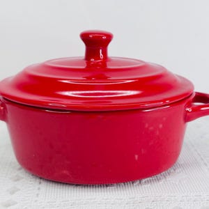 May include: A small, red ceramic casserole dish with a matching lid. The dish has two side handles and a small knob on the lid. The glossy finish reflects light, highlighting the vibrant red color. This item is suitable for baking and serving.