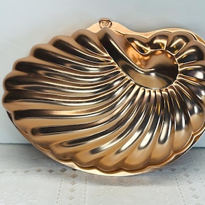 May include: A metallic copper-colored baking mold shaped like a nautilus shell. The mold features a series of curved, raised ridges that spiral toward the center, creating a distinct shell pattern with a smooth, reflective finish.
