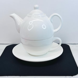 May include: A white ceramic tea set, including a teapot with a lid, a teacup, and a saucer. The teapot has a curved spout and handle. The teacup sits inside the teapot, and the saucer is beneath. The set is on a black surface.