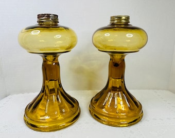 May include: Two amber glass oil lamps with fluted bases and clear glass chimneys. The lamps are on a white surface.