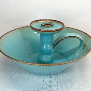 May include: A turquoise ceramic candle holder with a handle. The holder has a round base and a central column for the candle. The rim and the top of the candle holder are a darker brown color. The piece is on a white surface.