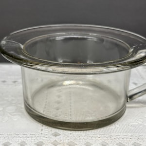 May include: A clear glass custard cup featuring a wide, flat rim and a single side handle. The vessel has a cylindrical body with a smooth, transparent finish, designed with a sturdy base and a uniform wall thickness throughout the entire piece.