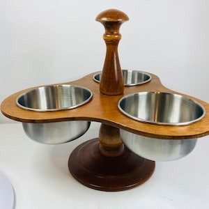 May include: A wooden condiment holder with three stainless steel bowls. The holder has a brown wood finish and a round base. The bowls are attached to the holder with a wooden post.