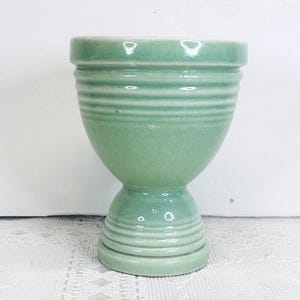 May include: A light green ceramic egg cup with a pedestal base. The egg cup has a rounded bowl shape with horizontal ridged detailing around the top and base. The cup is sitting on a white lace tablecloth.