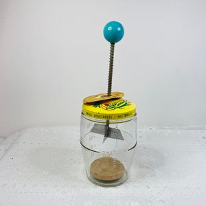 May include: Vintage glass food chopper with a yellow lid featuring text and a vegetable illustration. It has a metal blade, a wooden base, and a turquoise ball handle. The jar is marked with measurements.