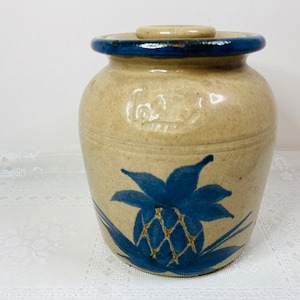 May include: Tan ceramic jar with a blue pineapple design. The jar has a lid and a dark blue rim. The pineapple design is centered on the front of the jar. The jar has a rustic, handmade appearance.