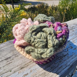 Pode incluir: Um vaso de suculentas de crochê com flores rosa, verde e roxas. O vaso é feito de fio e tem uma base bege.