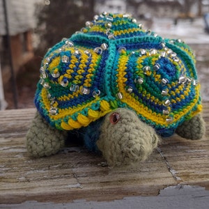May include: A crocheted turtle toy with a green, blue, and yellow shell. The shell is decorated with silver beads. The turtle has a brown eye and a green head.