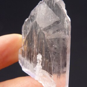 May include: A translucent, crystalline mineral specimen with a pale, almost colorless appearance. The crystal has a sharp, angular form with visible striations and a slightly rough texture. The background is a solid black.