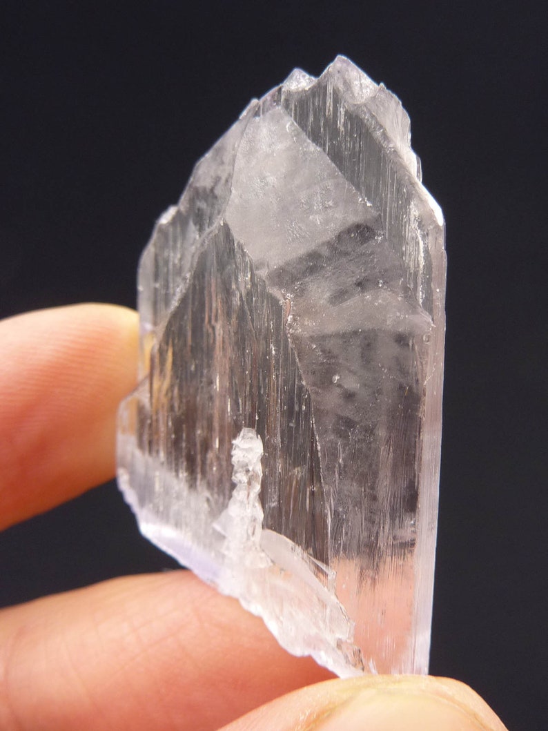 May include: A translucent, crystalline mineral specimen with a pale, almost colorless appearance. The crystal has a sharp, angular form with visible striations and a slightly rough texture. The background is a solid black.