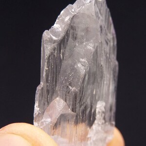 May include: A translucent, crystalline mineral specimen with a jagged edge. The crystal is held between fingers, showcasing its pale, almost colorless appearance. The mineral's surface has vertical striations, and the background is dark.