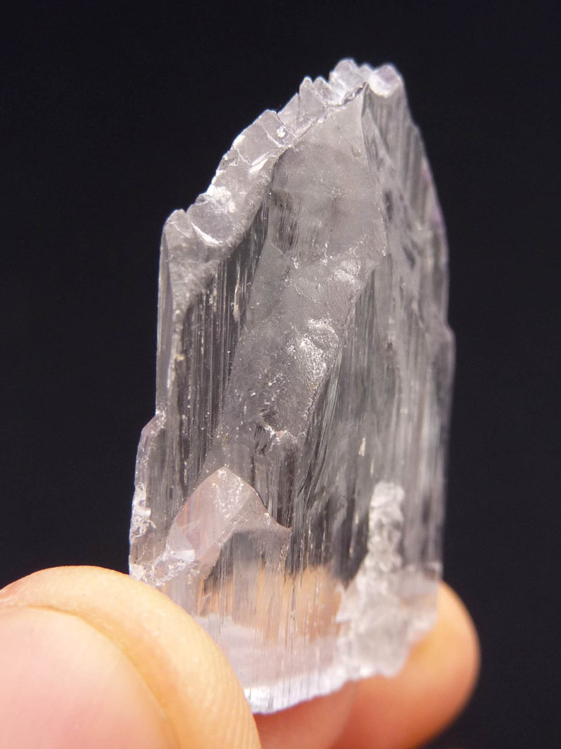 May include: A translucent, crystalline mineral specimen with a jagged edge. The crystal is held between fingers, showcasing its pale, almost colorless appearance. The mineral's surface has vertical striations, and the background is dark.