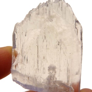 May include: A translucent, light-colored crystal with a jagged top edge and visible internal striations. The crystal is held between fingers, showcasing its natural, raw form. The overall color is a pale, almost colorless, with subtle variations in tone.