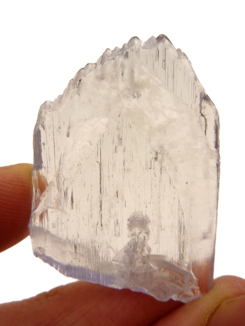 May include: A translucent, light-colored crystal with a jagged top edge and visible internal striations. The crystal is held between fingers, showcasing its natural, raw form. The overall color is a pale, almost colorless, with subtle variations in tone.