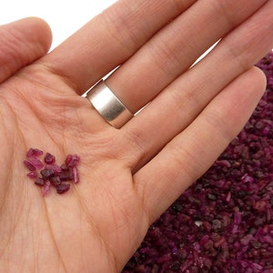 Puede incluir: Primer plano de una mano que sostiene una pequeña colección de piedras preciosas de rubí en bruto. Las piedras preciosas son de varios tonos de rojo y rosa. Un montón de piedras similares está en el fondo. Se ve un anillo de plata.