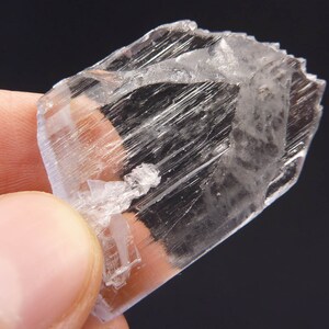 May include: A clear, crystalline mineral specimen with a rectangular shape. The mineral has a translucent appearance with visible internal striations and a slightly rough texture. The edges are sharp and well-defined, and the overall color is a pale, almost colorless, white.
