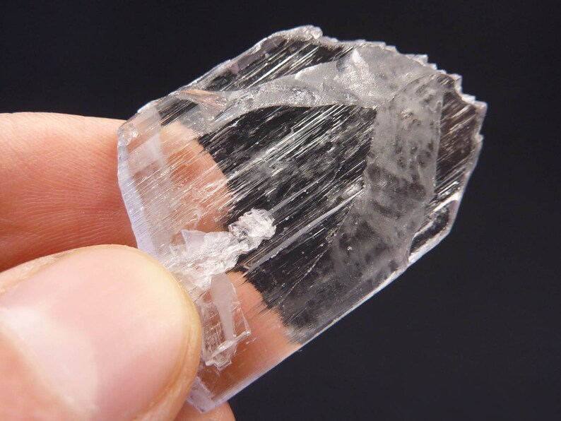 May include: A clear, crystalline mineral specimen with a rectangular shape. The mineral has a translucent appearance with visible internal striations and a slightly rough texture. The edges are sharp and well-defined, and the overall color is a pale, almost colorless, white.