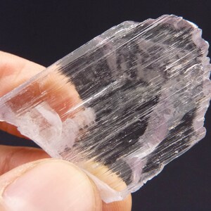 May include: A translucent, crystalline mineral specimen with a pale lavender hue. The mineral has a layered structure with visible striations, held by a person's fingers against a dark background. The edges are rough and uneven.