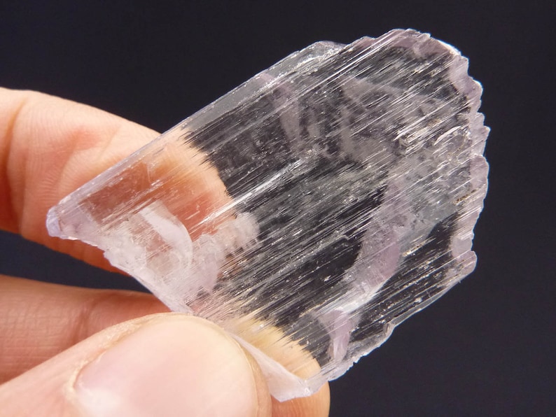 May include: A translucent, crystalline mineral specimen with a pale lavender hue. The mineral has a layered structure with visible striations, held by a person's fingers against a dark background. The edges are rough and uneven.