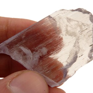May include: A translucent, light purple selenite crystal with a layered, fibrous texture. The crystal is held in a hand against a white background, showcasing its natural, raw form. The mineral has a slightly iridescent quality.
