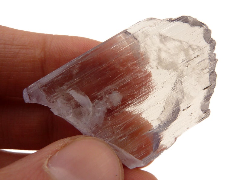 May include: A translucent, light purple selenite crystal with a layered, fibrous texture. The crystal is held in a hand against a white background, showcasing its natural, raw form. The mineral has a slightly iridescent quality.