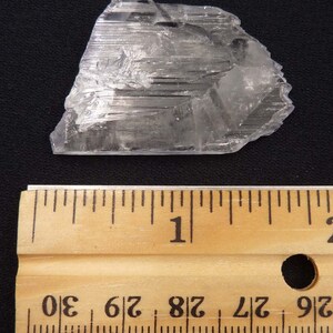 May include: A clear, crystalline mineral specimen with a layered structure. The mineral is positioned next to a wooden ruler, with measurements in inches. The ruler shows measurements up to 30 inches.