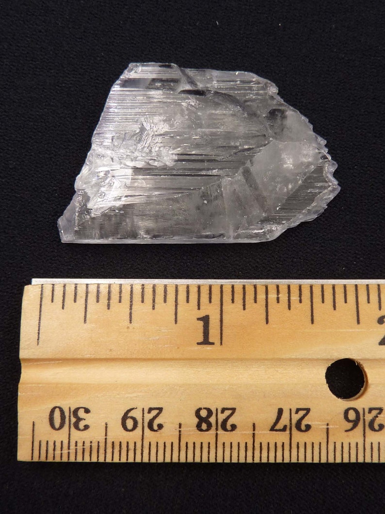 May include: A clear, crystalline mineral specimen with a layered structure. The mineral is positioned next to a wooden ruler, with measurements in inches. The ruler shows measurements up to 30 inches.