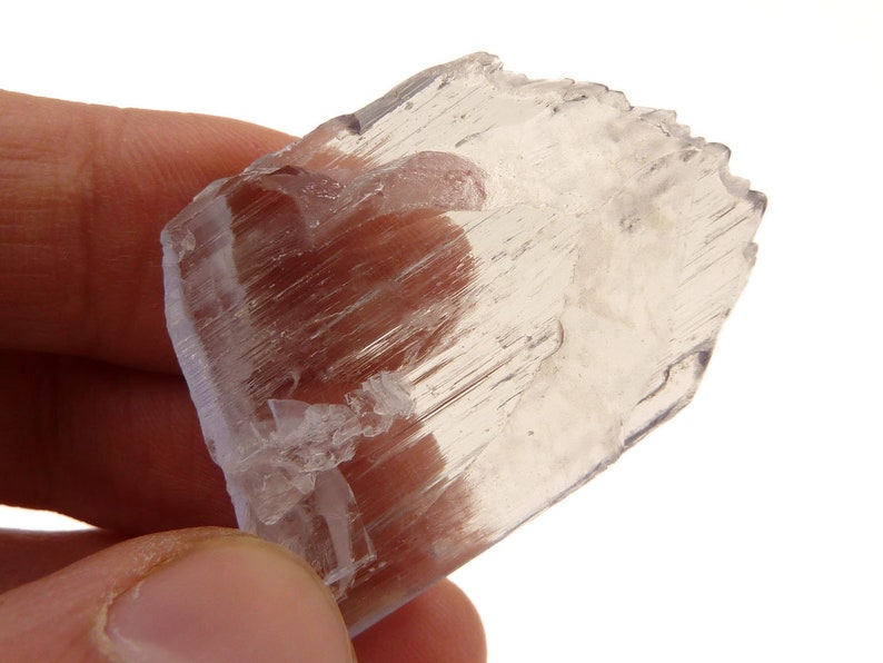 May include: A translucent, light-colored selenite crystal with a layered, fibrous structure. The crystal has a rough, uneven edge and a slightly curved shape. The background is white.