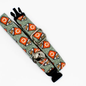 May include: A green and orange cat collar with a bell and a buckle closure. The collar has a pattern of orange boxes with a white bone inside.