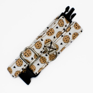 May include: A white cat collar with a brown cookie pattern and a silver bell. The collar has a black adjustable buckle.