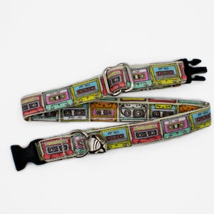 May include: A dog collar featuring a repeating pattern of colorful cassette tapes in shades of pink, blue, yellow, and green. The collar has a silver metal buckle and a black plastic clasp. The collar is approximately 1 inch wide.