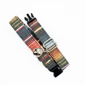 May include: Two colorful cat collars with a bell. The collars are made of fabric with a striped pattern in shades of red, orange, blue, and gray. The collars have a black buckle and a silver bell.
