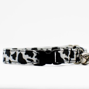 Peut inclure: Un collier de chat imprim&eacute; vache noir et blanc avec une boucle noire et une clochette argent&eacute;e.