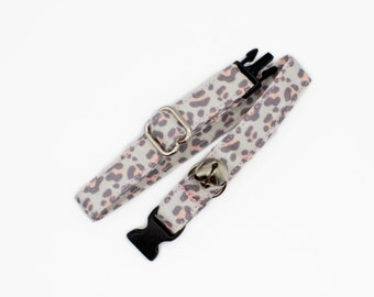 May include: A gray and pink leopard print cat collar with a black buckle and a silver bell.