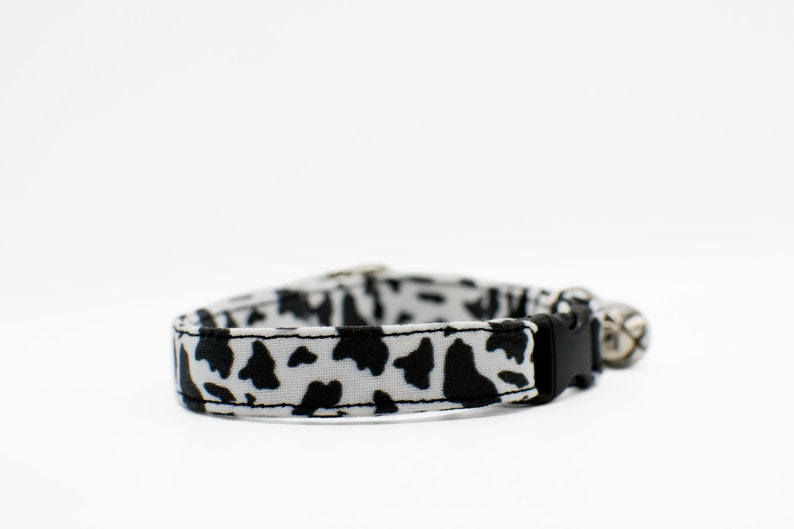 Peut inclure: Un collier de chat imprim&eacute; vache noir et blanc avec une boucle noire et une clochette argent&eacute;e.
