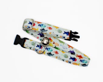 May include: Two cat collars with a colorful fish pattern on a light blue background. One collar has a silver bell and a metal loop, while the other has a black plastic buckle. The collars are designed for pets.