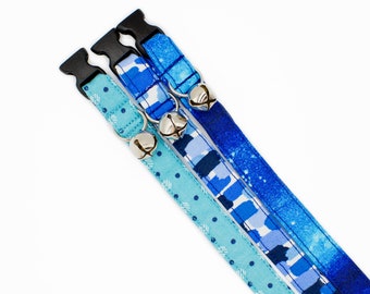 May include: Three blue cat collars with bells. The collars are made of fabric with different patterns: polka dots, abstract brushstrokes, and a watercolor-like design.