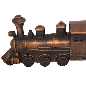 May include: A detailed bronze-coloured toy train with a classic locomotive design. The train features a smokestack, multiple wheels, and a cabin with windows. The overall aesthetic is vintage, suggesting a collectible or decorative item.