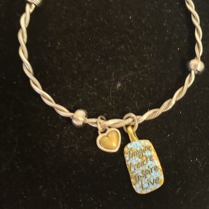 Puede incluir: Pulsera de brazalete plateada con un dise&ntilde;o de cuerda trenzada. La pulsera presenta un dije de coraz&oacute;n dorado y un dije rectangular con las palabras "Imagine Create Inspire Live" en negro y azul.