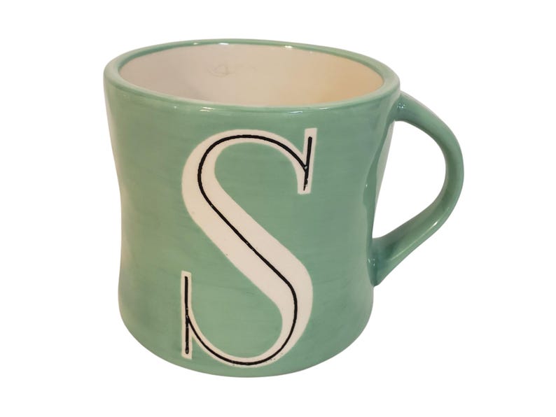 May include: A ceramic mug with a light teal exterior and a white interior. The mug features a large white letter "S" with a black outline. The handle is curved and the mug has a slightly curved shape.