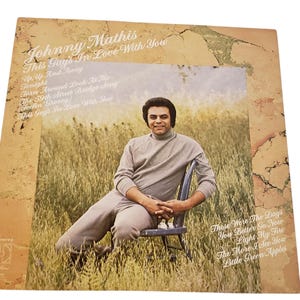 May include: Vintage vinyl record album cover featuring Johnny Mathis. The album is titled "This Guy's In Love With You." The cover art shows Mathis seated in a chair in a field. The album's song titles are listed on the cover.