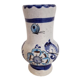 May include: A small, white ceramic vase with blue floral and butterfly designs. The vase has a flared top with a scalloped edge, a blue band around the neck, and a rounded base. The vase is approximately 6 inches tall.