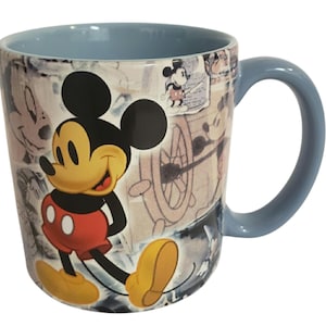 May include: A ceramic mug featuring Mickey Mouse. The mug has a light blue interior and handle. The exterior showcases a collage of Mickey Mouse sketches and illustrations, with a large image of Mickey in his classic outfit.
